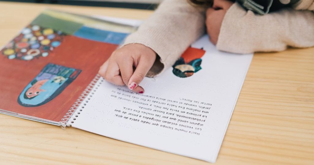 Montessori Literacy and the Science of Reading: Insights from Zil Jaeger. Image shows a wirebound book laying open on a wooden table. A child's arms are seen with a finger tracking the words on the page.
