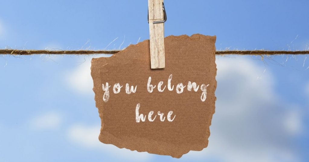 A brown paper sign hangs on a string with blue sky in the background The sign reads "you belong here." The picture is intended to promote diversity and inclusion.
