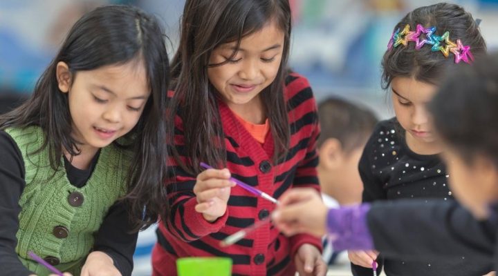 Human Tendencies as Witnessed in the Elementary Child 
Image shows three smiling elementary aged students in a classroom standing at a table and painting. The student on the left is wearing a green sweater vest with three buttons. The student in the middle is wearing a red sweater with black striped. The student on the right is wearing a black shirt with a coloring headband in her hair. All three students are holding purple paint brushes. 