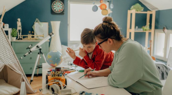 A Quick Guide to Montessori Homeschool. 
Image shows a parent helping a child with school assignment in the foreground. They are sitting at a desk writing in a notebook. In the background is the child's bedroom, painted in soft green and blue colors with a telescope and a globe. 