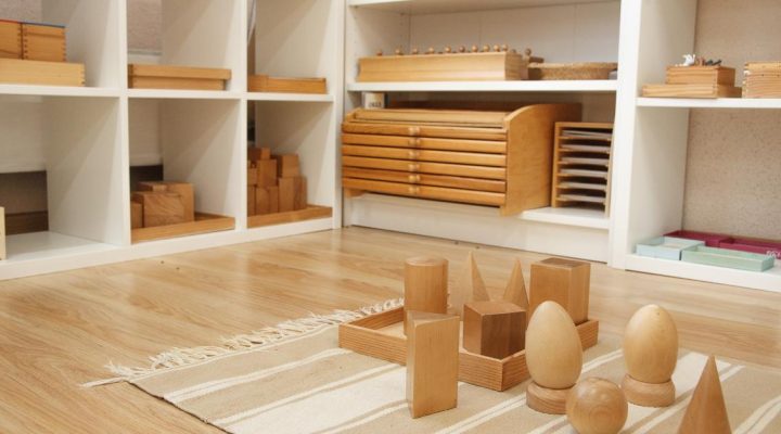 Virtual Homeschooling. Virtual homeschooling with Montessori. Image shows white shelves with various wooden Montessori sensorial materials in the background. In the foreground, there is a tan and white rug with several wooden geometric solids displayed.