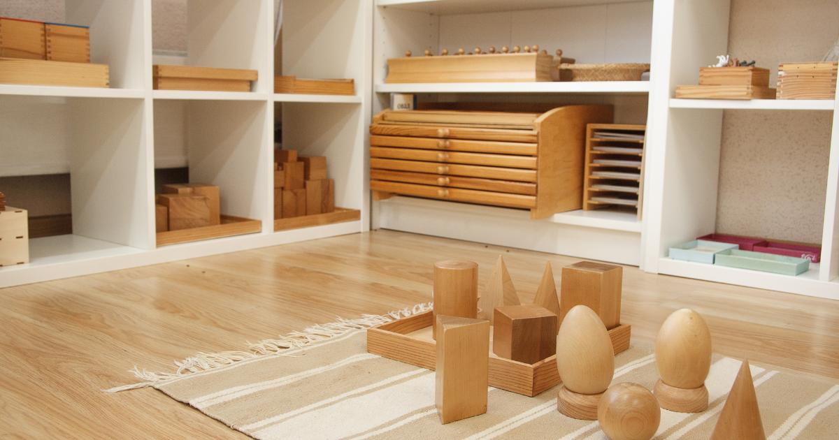 Virtual homeschooling with Montessori. Image shows white shelves with various wooden Montessori sensorial materials in the background. In the foreground, there is a tan and white rug with several wooden geometric solids displayed.