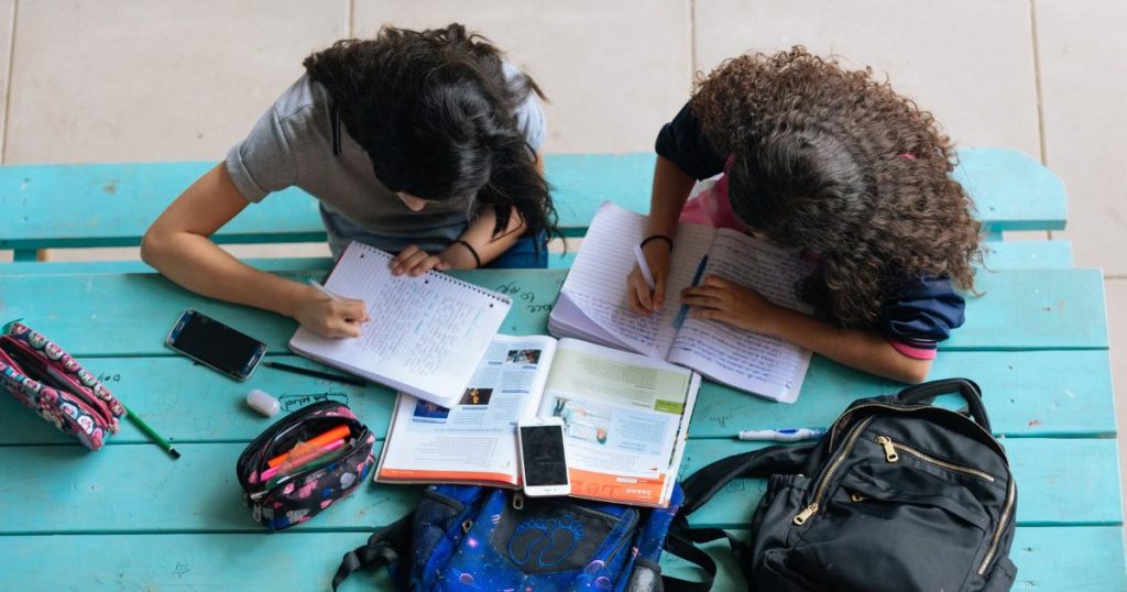 When you have a small group of older children in your class. Image shows two students with long dark hair sitting side by side at blue table. Both students have their heads down writing in their notebooks. Also on the table are backpacks, an open textbook, cell phones, and pencil cases.
