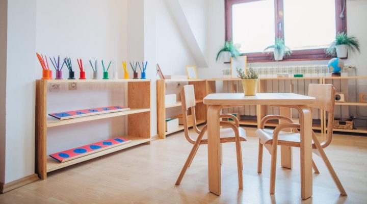 Embracing Bilingual Education in Montessori Schools. Image shows a bright and tranquil Montessori classroom that is ready for students. In the foreground there is a wooden table with two chairs. The metal insets with colored pencils can be seen in the background. There is a large window providing sunlight with plants scattered around the room. 