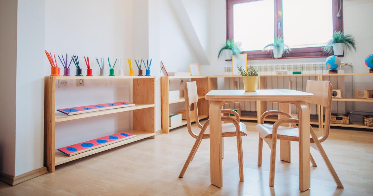 Embracing Bilingual Education in Montessori Schools. Image shows a bright and tranquil Montessori classroom that is ready for students. In the foreground there is a wooden table with two chairs. The metal insets with colored pencils can be seen in the background. There is a large window providing sunlight with plants scattered around the room.