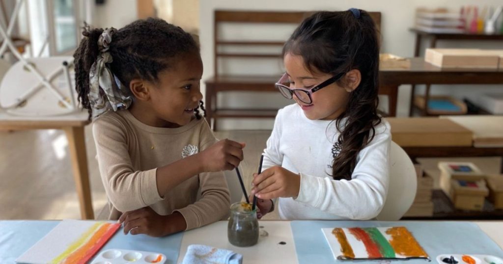Dahlia Montessori demonstrates the power of Montessori education, multilingual immersion, and community-focused learning. Image shows two children smiling and holding paintbrushes dipped in a paint jar.