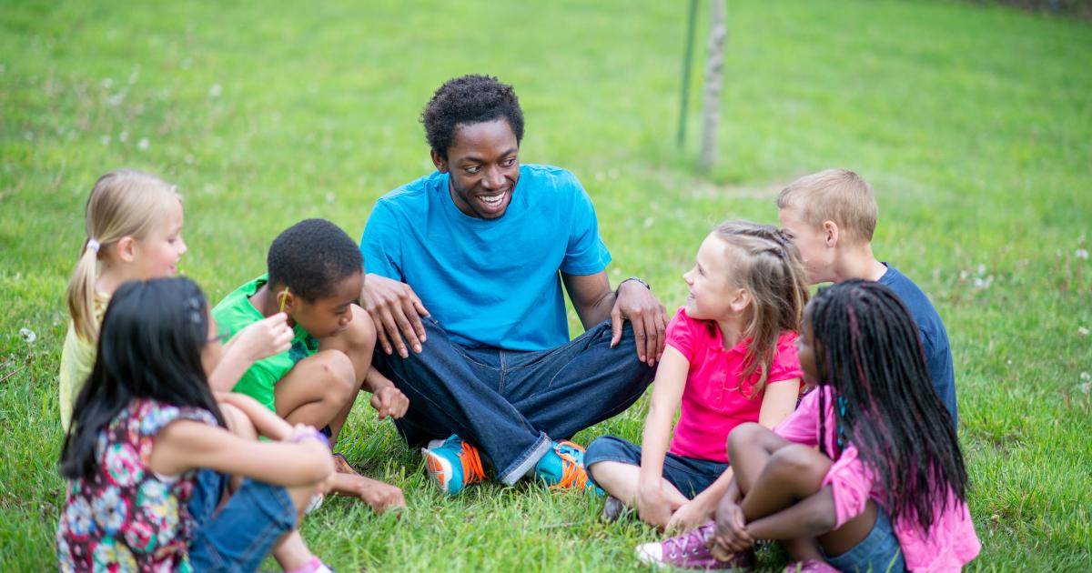 Bringing Joy and Levity to Montessori Spaces. In this week's episode of the podcast, Letty is talking to Lisa Thauvette, former head of International Montessori School, and a speaker, trainer, and educator. Lisa shares why we should bring joy and levity into Montessori education. Image shows a group of children sitting in a circle outside in the grass, with their teacher sitting the middle. They are laughing and smiling at one another.