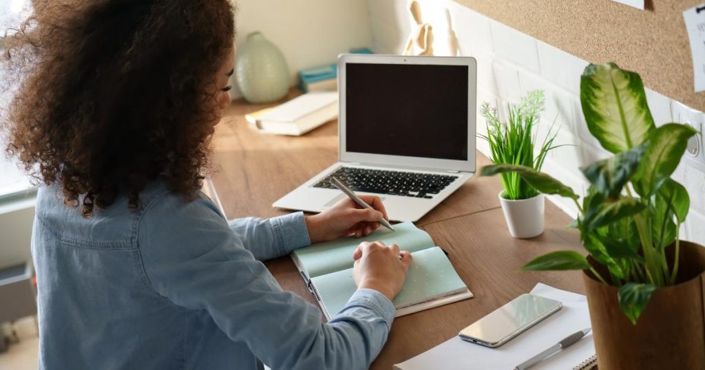 In the latest podcast episode, we are looking at planning and record keeping and discussing how both are essential for Montessori educators. Image shows a teacher sitting at a table writing in a notebook with a pencil. There is a plant in the foreground and an open laptop in the background.