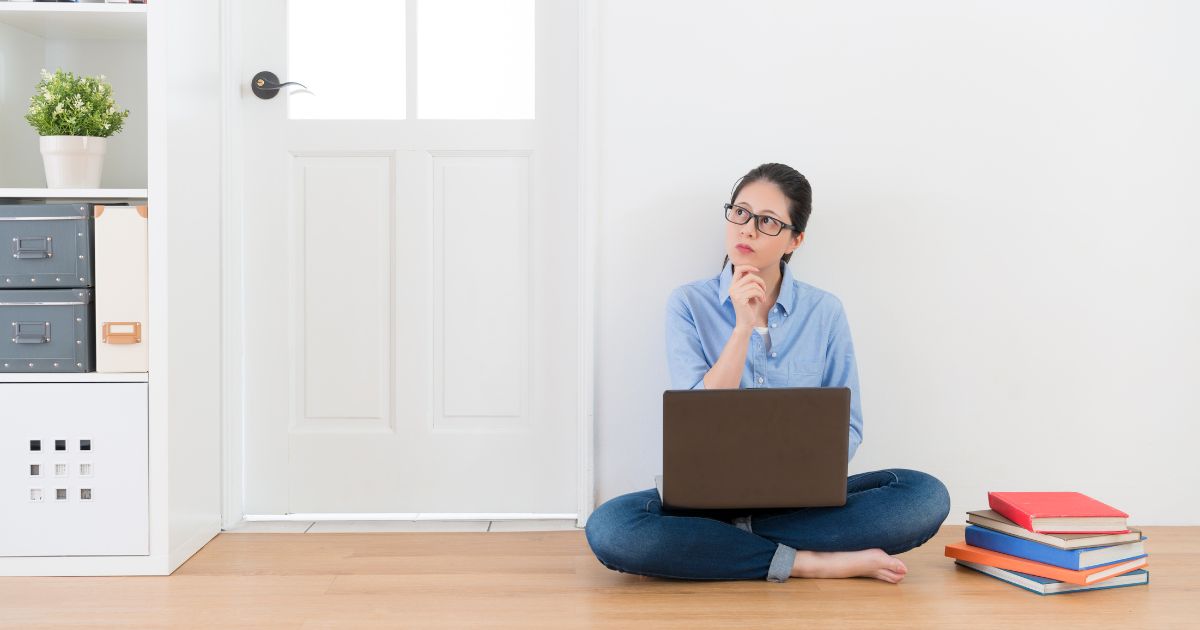 Elementary planning and record keeping. Image shows woman with glasses sitting cross legged on a wooden floor. She has a hand to her chin and a contemplative expression. An open laptop is on her lap and a stack of books is beside her.