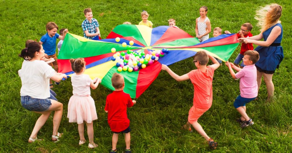 Building relationships through games. Image shows a small group of children and a teacher on the grass. They are in a circle holding up a multicolored parachute with colorful balls.
